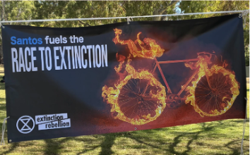 A photo of a banner that reads "Santos fuels the race to extinction" with an XR logo and a bicycle on fire.