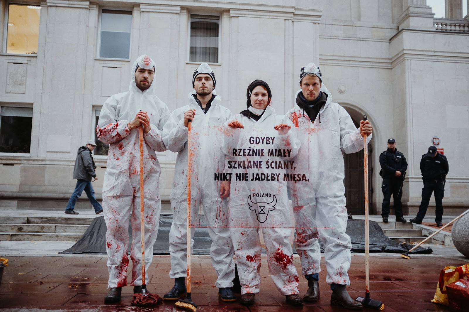 Four people wearing blood-covered plastic overalls hold bloody brooms